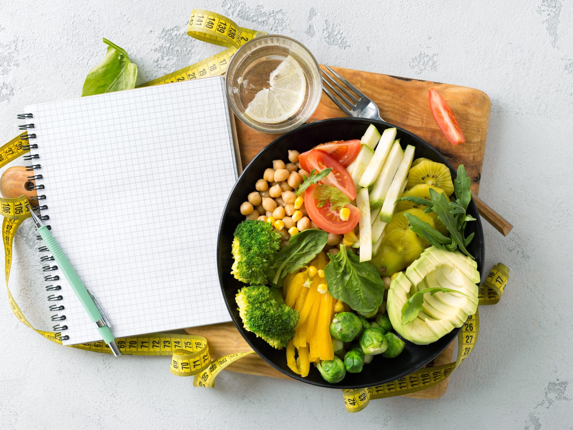 Vegan healthy balanced diet concept. Vegetarian buddha bowl with blank notebook and measuring tape. Сhickpeas, broccoli, pepper, tomato, spinach, arugula and avocado in plate on white background. Top view